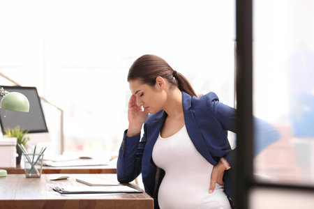 Young pregnant woman suffering from pain while working in officeの写真素材