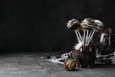 Yummy cake pops coated with chocolate in glass jar full of coffee beans on table. Space for text
