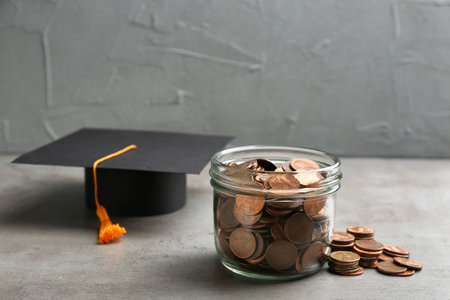 Glass jar, coins and graduation hat on gray table. Space for textの写真素材