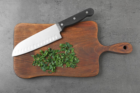 Wooden board with sharp knife and chopped parsley on gray background, top view