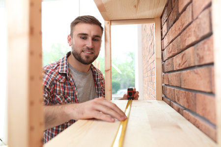 Young working man measuring wooden shelf indoorsの写真素材