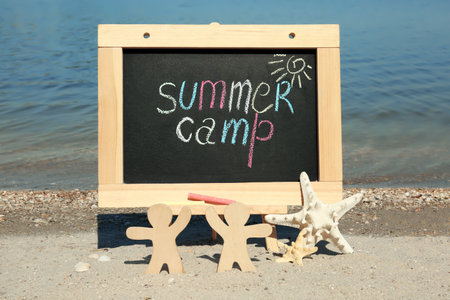 Small chalkboard with text SUMMER CAMP, wooden people and starfish on beach near riverの写真素材