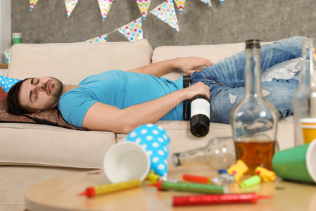 Young man with bottle of wine sleeping on sofa in messy room after partyの写真素材