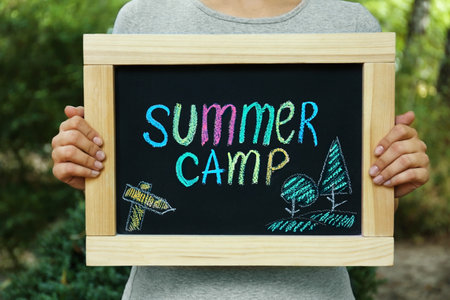 Woman holding blackboard with text SUMMER CAMP and drawing on blurred background, closeupの写真素材