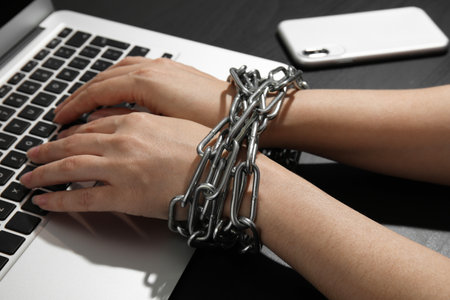Woman with chained hands using laptop on black background, closeup. Loneliness conceptの写真素材