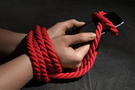Woman with roped hands holding smart phone on gray background, closeup. Solitude concept