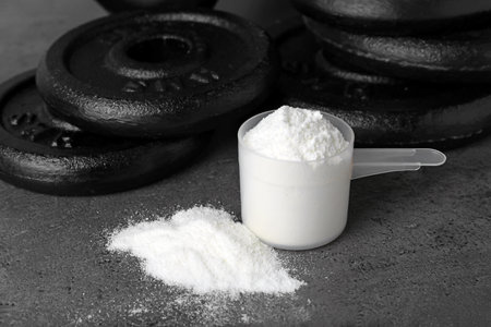 Protein powder, measuring scoop and dumbbell plates on gray table. Space for text