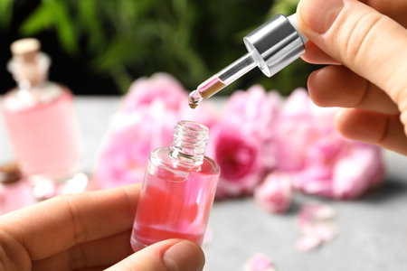 Woman dripping rose essential oil into bottle over table, closeup