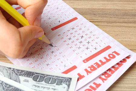 Woman filling out lottery tickets with pencil and money on wooden table, closeup. Space for textの写真素材