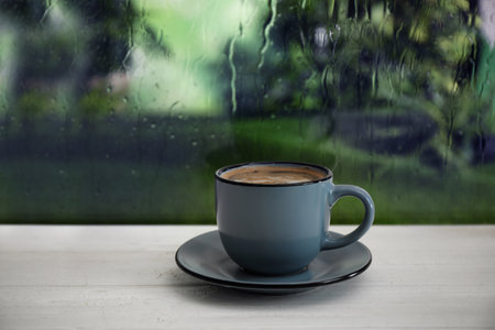 Cup of hot drink on wooden windowsill against glass with rain drops, space for textの写真素材