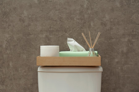 Tray with roll of paper, napkins and reed air freshener on toilet tank near gray wall. bathroom...