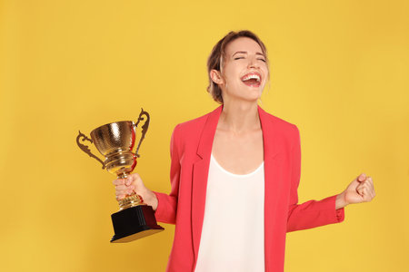 Portrait of happy young businesswoman with gold trophy cup on yellow backgroundの写真素材