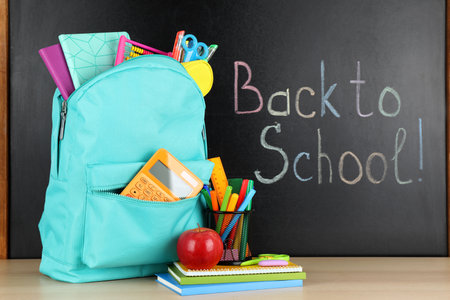 Bright backpack with school stationery on brown wooden table near black chalkboard. back to schoolの写真素材