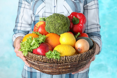 Woman holding wicker bowl with ripe fruits and vegetables on color background, closeupの写真素材