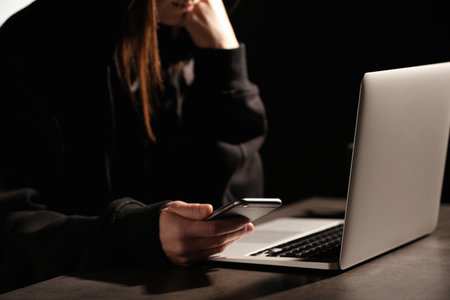 Woman using smartphone at table with laptop in darkness, closeup. Loneliness concept