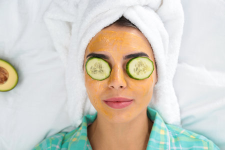 Young woman with facial mask and cucumber slices lying on bed, top viewの写真素材
