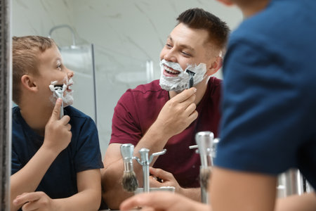 Dad shaving and son imitating him at mirror in bathroomの写真素材