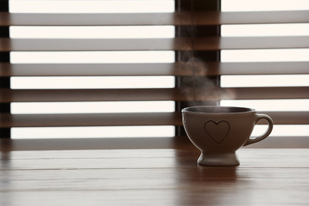 Cup of hot tea on wooden table near window with blinds. Space for textの写真素材
