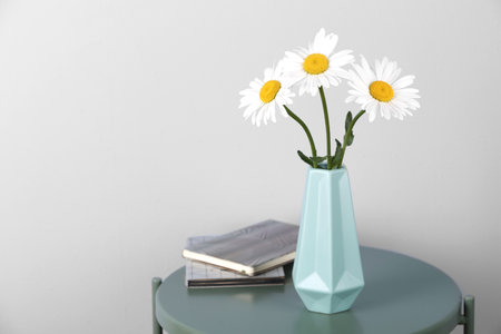 Beautiful tender chamomile flowers in vase and notebooks on table against light background, space for textの写真素材