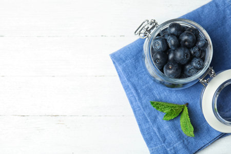 Glass jar of tasty fresh blueberries, mint leaves and fabric on white wooden table, flat lay with space for textの写真素材