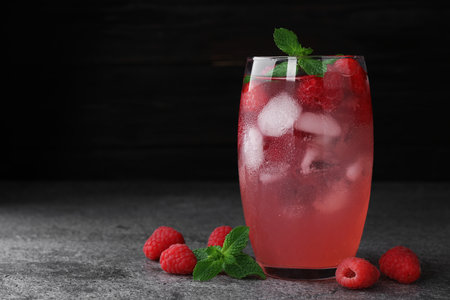 Glass of refreshing drink with raspberry and mint on gray stone table against dark background, space...