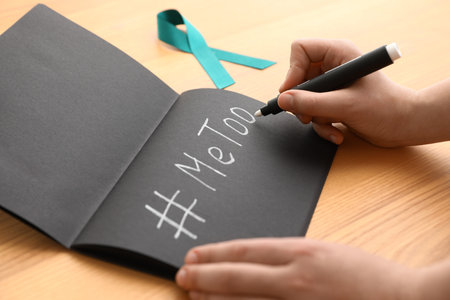 Woman writing hashtag MeToo in notebook near teal awareness ribbon at table, closeup. Stop sexual assaultの写真素材