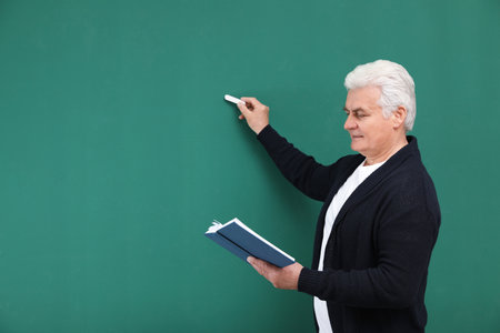 Portrait of senior teacher with book and chalk at green board, space for textの写真素材