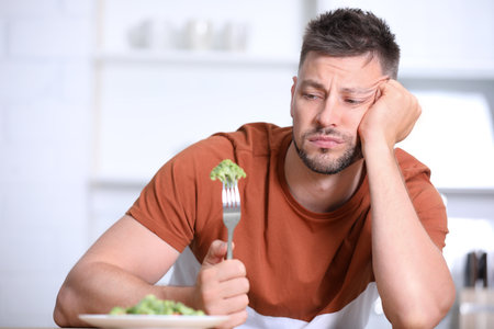 Portrait of unhappy man eating broccoli salad in kitchenの写真素材
