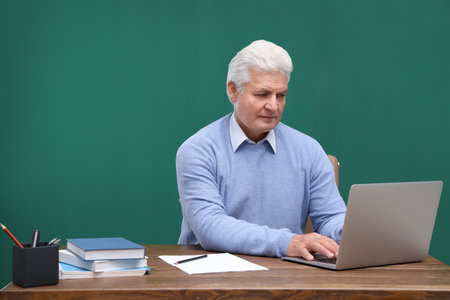 Portrait of senior teacher with laptop at table against green chalkboardの写真素材