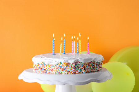 Tasty birthday cake with burning candles and balloons on orange backgroundの写真素材
