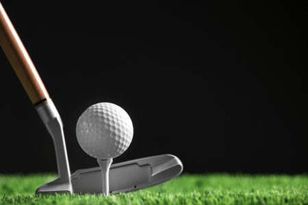 Hitting golf ball with club on artificial grass against black background, space for text