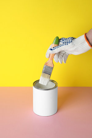 Person dipping brush into can of white paint on pink table against yellow background, closeup. Mockup for designの写真素材