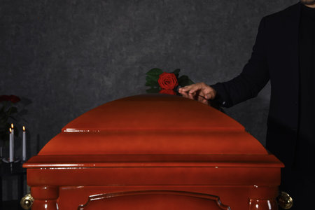 Young man near casket with red rose in funeral home, closeup