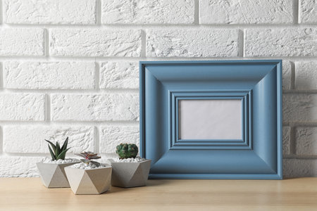 Blank frame and succulent plants on wooden table near white brick wall, space for design. home decor