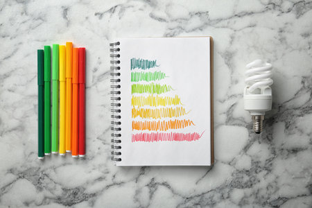 Notebook with colorful bars, markers and fluorescent light bulb on white marble background, flat...