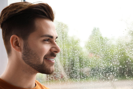 Smiling handsome man near window indoors on rainy dayの写真素材
