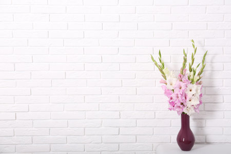 Vase with beautiful gladiolus flowers on table against white brick wall. Space for textの写真素材