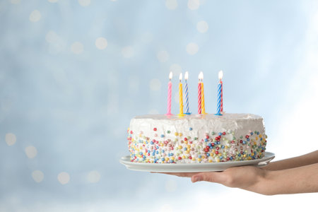 Woman holding birthday cake with burning candles against blurred background, closeup. Space for textの写真素材