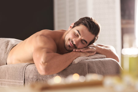 Handsome young man relaxing on massage table in spa salonの写真素材