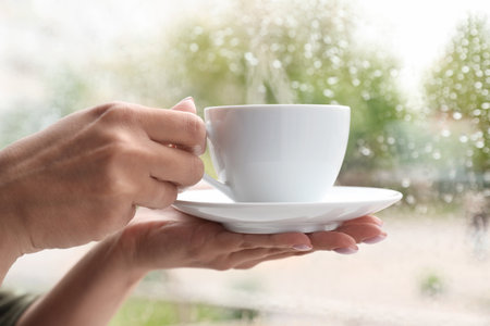 Young woman with cup of coffee near window indoors on rainy day, closeupの写真素材