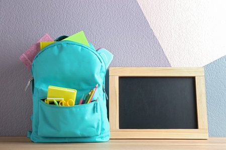Backpack full of different school stationery and small blank chalkboard on table near color wall. Space for textの写真素材