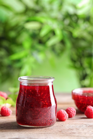 Glass jar of sweet jam with ripe raspberries on wooden table against blurred background