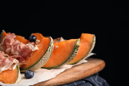 Wooden board with melon, prosciutto and blueberries on table against black backgroundの写真素材