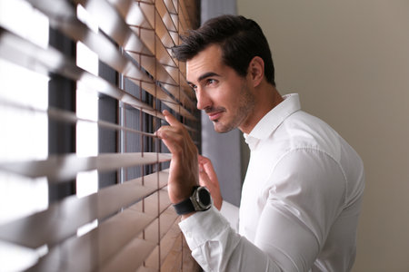Handsome young man looking through window blinds indoorsの写真素材