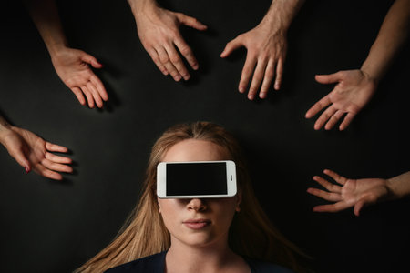 Young woman with smartphone covering her eyes surrouded by people's hands on black background, top view. Space for textの写真素材