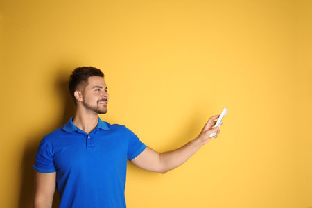 Happy young man operating air conditioner with remote control on yellow background. Space for textの写真素材