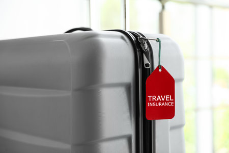 Stylish suitcase with travel insurance label on blurred background, closeup. Space for textの写真素材