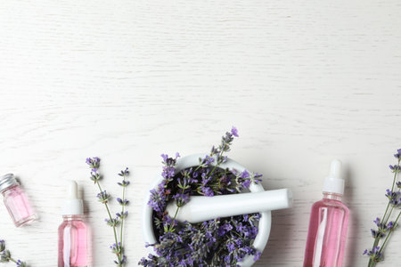 Bottles of essential oil, mortar and pestle with lavender flowers on white wooden background, flat...