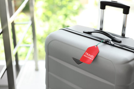 Stylish suitcase with travel insurance label on blurred background, closeup. Space for textの写真素材