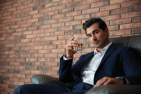 Young man with glass of whiskey near brick wall indoors. Space for textの写真素材